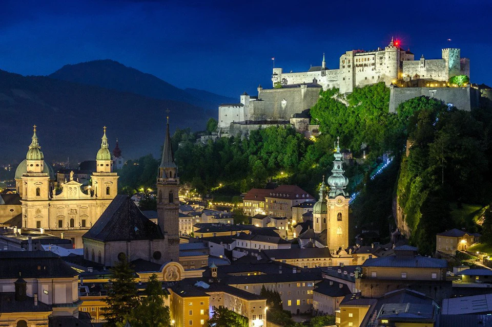 A brightly lit historic castle atop a hill, overlooking a town's church steeples and buildings at night with mountains in the background.