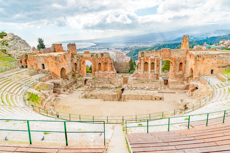 Ancient stone amphitheater ruins with tiered seating and scenic views of the coastline and mountains in the background.
