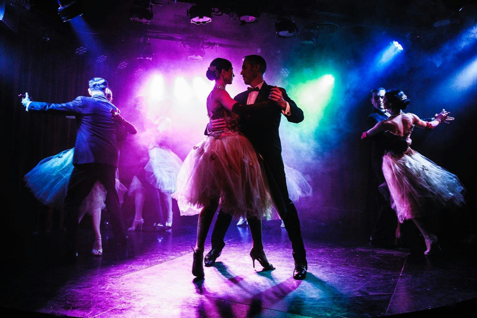 A couple in formal attire dancing under colorful stage lights, surrounded by other dancing couples in a dimly lit space.