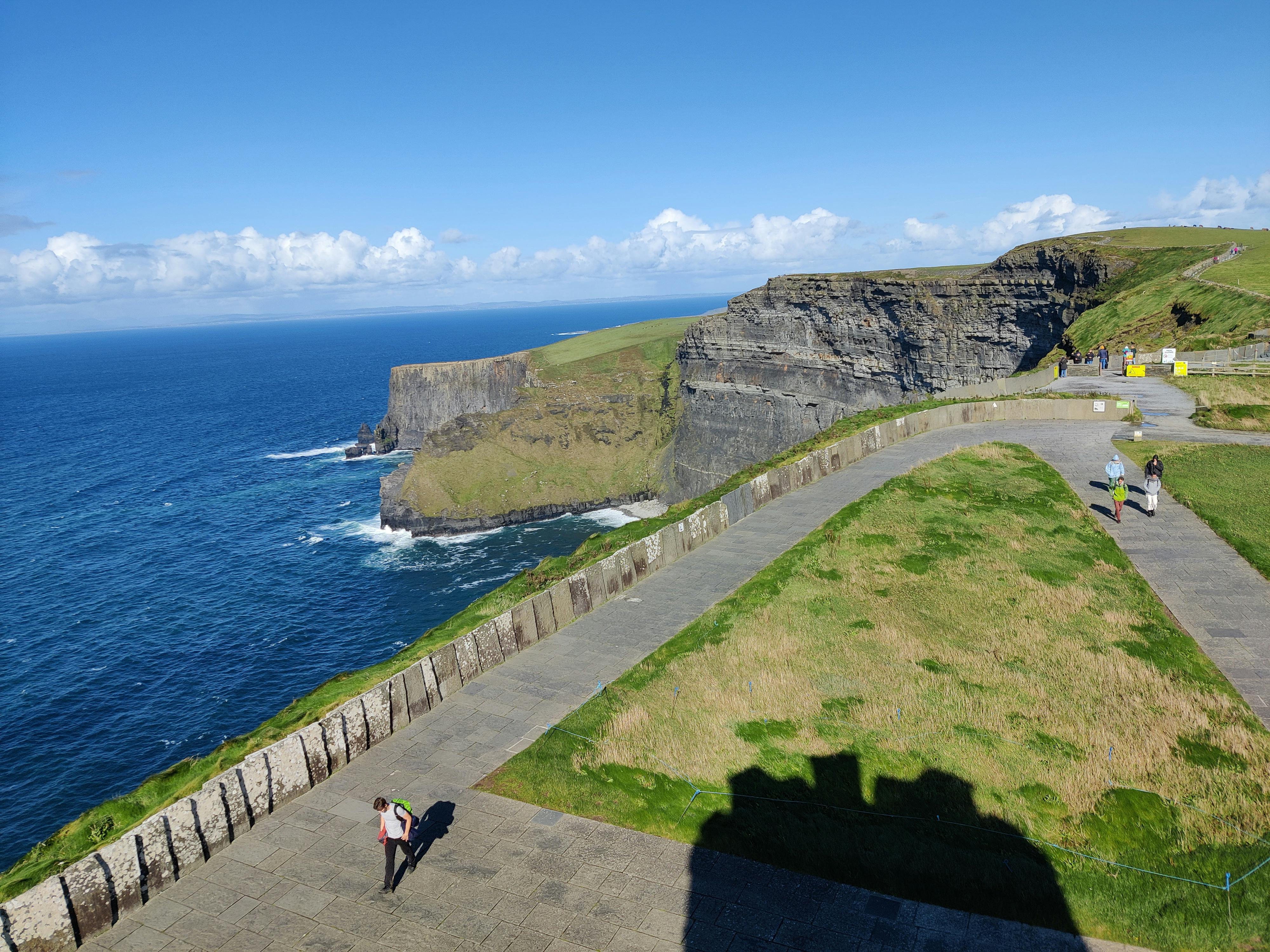 Person standing on a walkway near grassy cliffs overlooking blue ocean under clear sky.