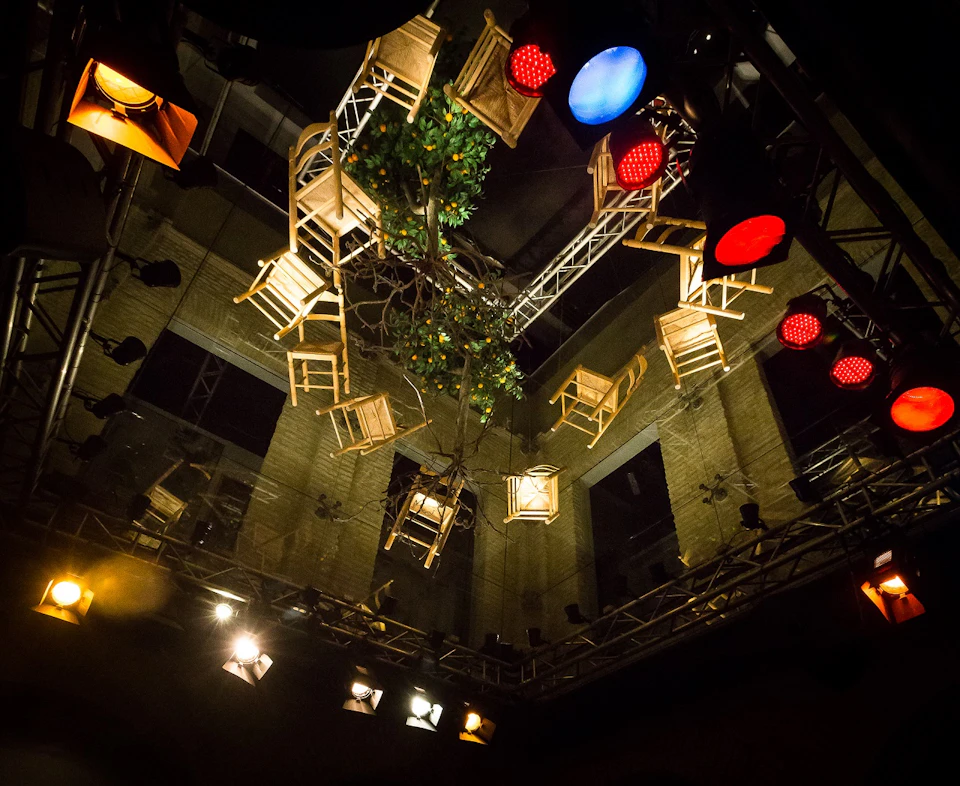 Wooden chairs and a plant suspended from the ceiling amidst stage lights, with red, blue, and yellow spotlights illuminating them.