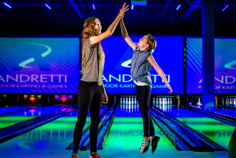 Two people high-fiving on a vibrant bowling lane with "Andretti Indoor Karting & Games" visible on screens in the background.