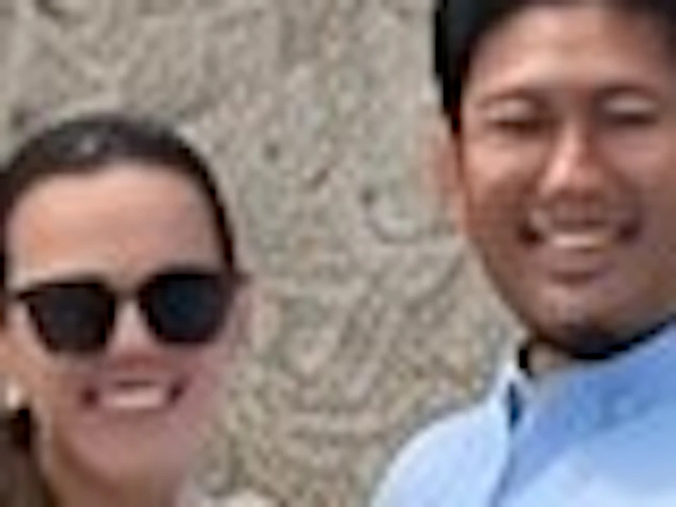 Two people pose in front of a large stone wall structure on a sunny day, with others visible on an upper walkway.