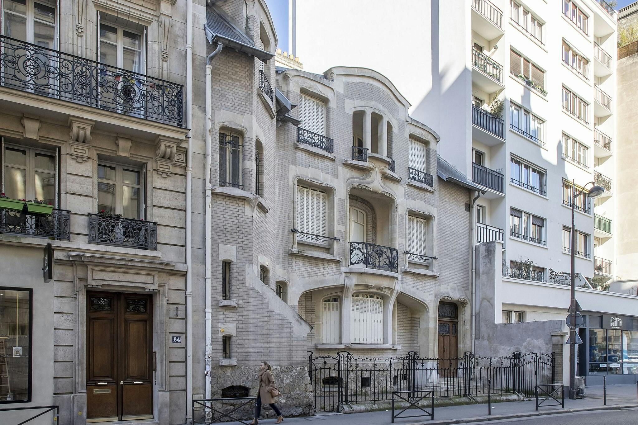 Edifici ornamentali a più piani con balconi intricati fiancheggiano una strada. Una persona cammina davanti a loro, vestita con un cappotto marrone.