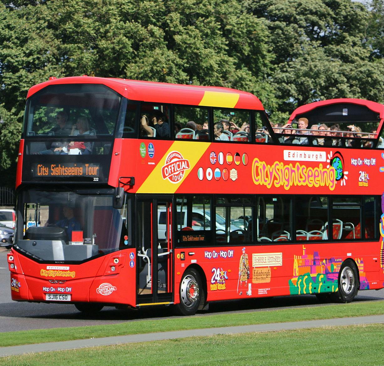 Autobus rosso brillante a due piani con tettuccio aperto, con la scritta "City Sightseeing", pieno di passeggeri, che viaggia in un parco.