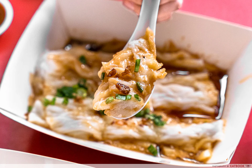 A close-up of a spoon holding a portion of rice noodles with sauce, garnished with green onions and bits of meat.