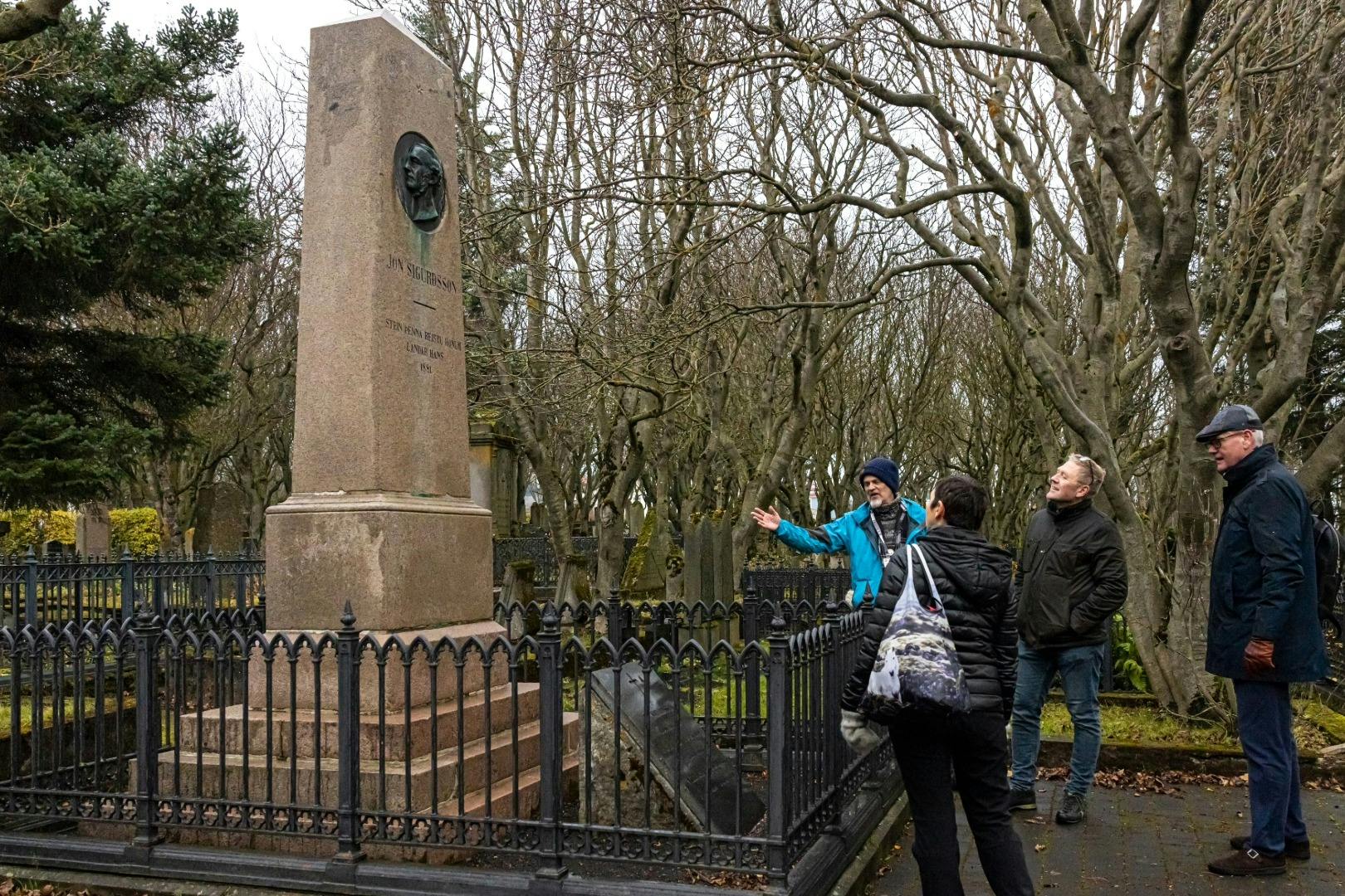 낮에는 나뭇잎이 없는 나무로 둘러싸인 묘지의 울타리 비석 근처에 세 사람이 서 있습니다.