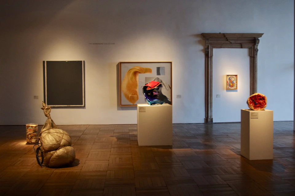 Gallery with modern art, including abstract paintings, a metallic sculpture on a plinth, and a golden cushioned piece.