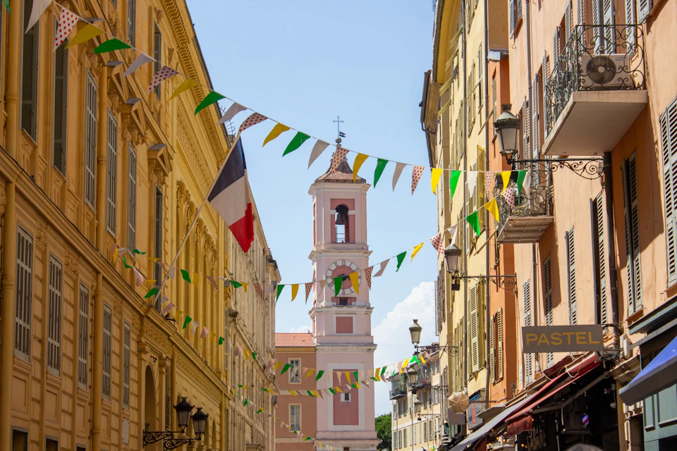 A narrow street decorated with colorful flags, featuring a pink bell tower and a French flag on a sunny day.