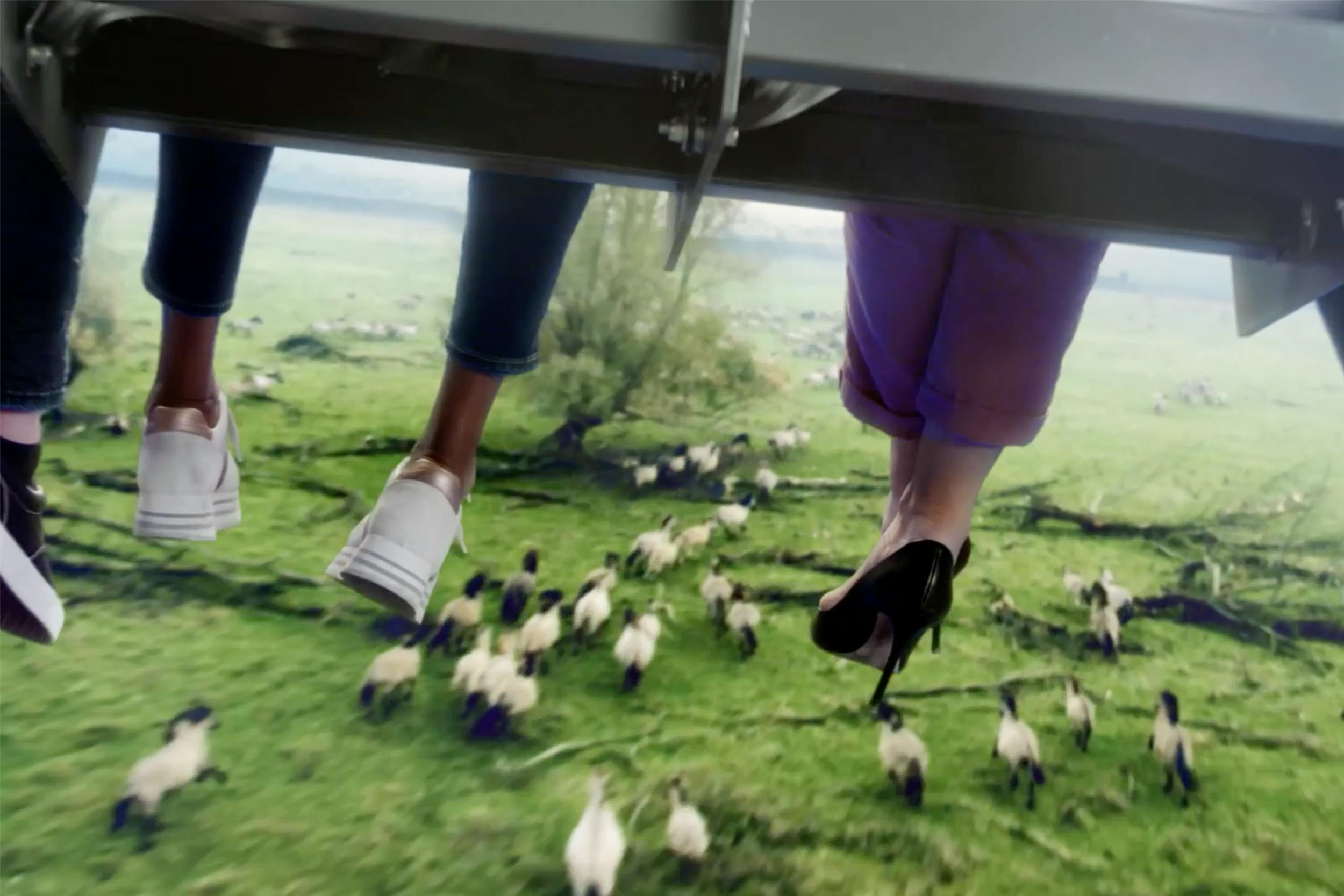 Δύο ζευγάρια πόδια κρέμονται πάνω από ένα χορταριασμένο χωράφι με πρόβατα. Ο ένας φοράει λευκά αθλητικά παπούτσια, ο άλλος μαύρα ψηλά τακούνια.