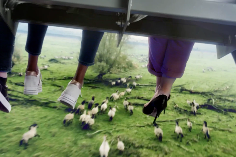 Two pairs of legs dangle above a grassy field with sheep. One wears white sneakers, the other black high heels.