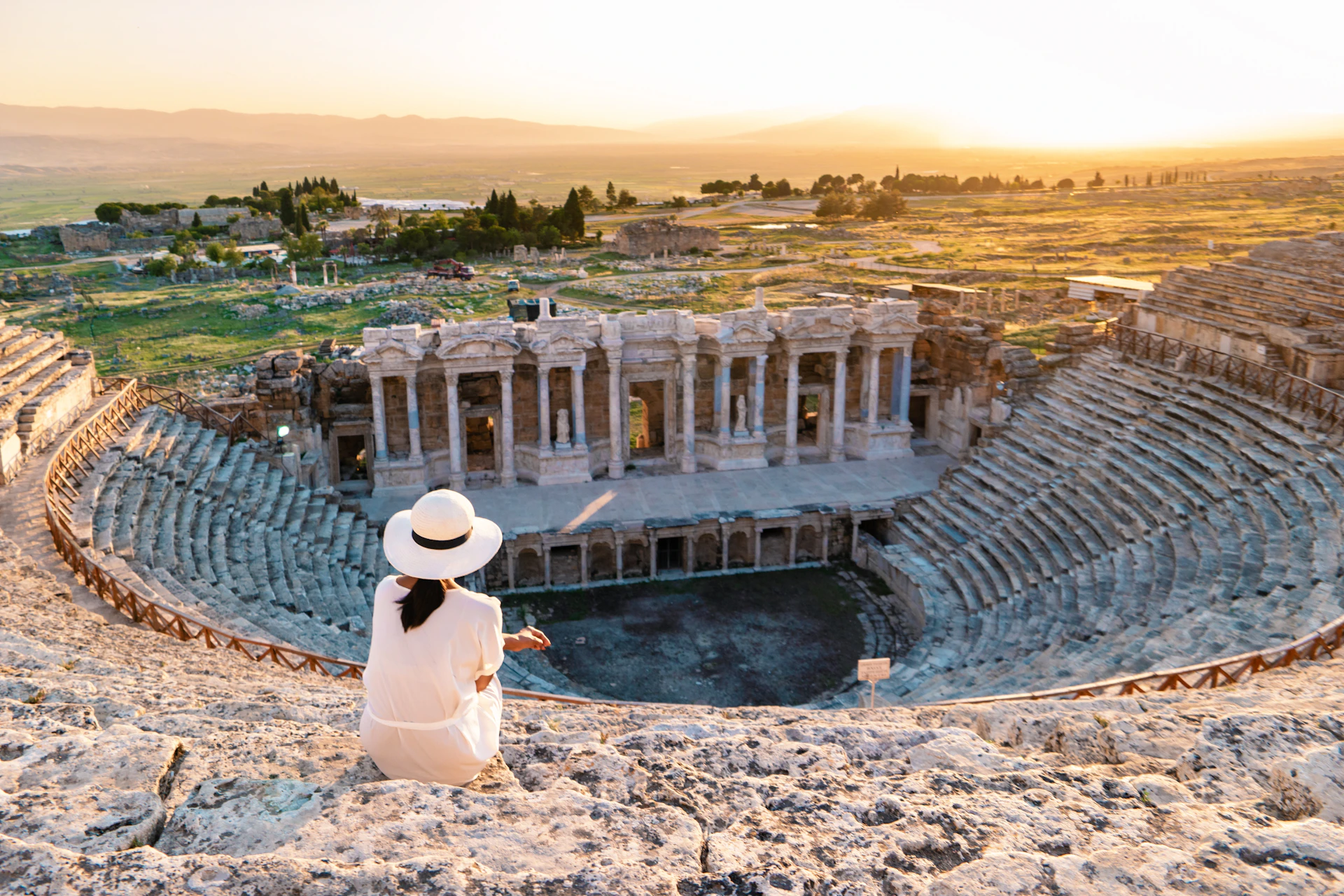 Person in white and hat sits atop an ancient amphitheater, overlooking ruins and a landscape bathed in sunset light.