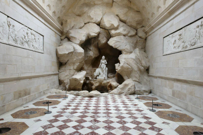 Statue of a woman and child in a rocky alcove at the end of a hall with a checkered floor in a stone-walled room.