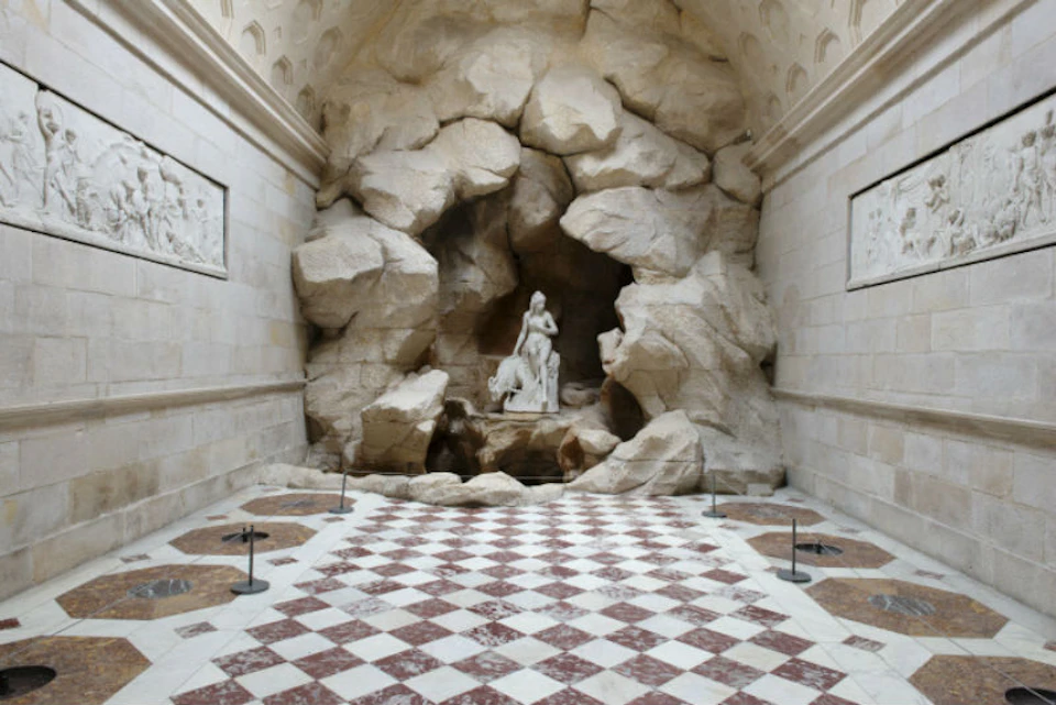Statue of a woman and child in a rocky alcove at the end of a hall with a checkered floor in a stone-walled room.