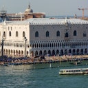 A large, ornate building stands next to a bustling waterfront in Venice, with a boat in the foreground and a dome in the background.