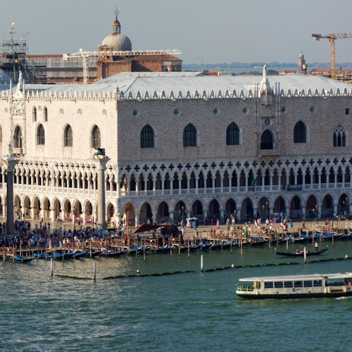A large, ornate building stands next to a bustling waterfront in Venice, with a boat in the foreground and a dome in the background.
