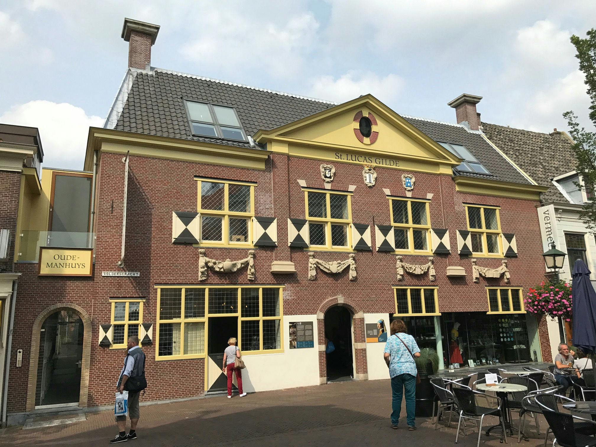 A historic brick building with yellow trim labeled &#34;St. Lucas Gilde&#34; and &#34;Oude Manhuys.&#34; People walk and sit at outdoor tables.