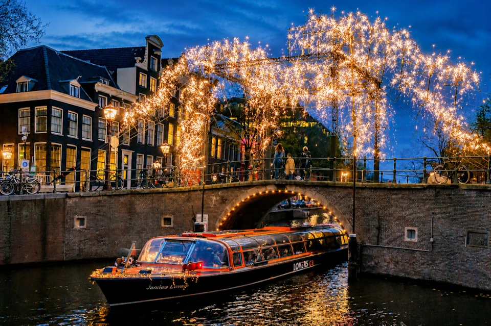 A canal boat passes under a bridge decorated with bright string lights during the evening in a city with traditional houses.