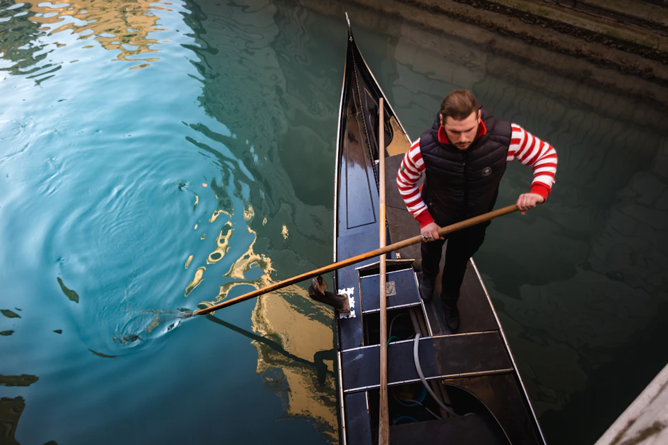 A person wearing a black vest and striped shirt is standing on a gondola, using a pole to navigate a calm waterway.