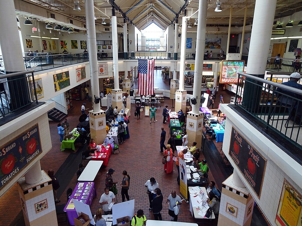 Marktszene in einer Halle mit Ständen und Tischen, Menschen, die spazieren gehen und sich unterhalten, in der Mitte hängt eine große amerikanische Flagge.