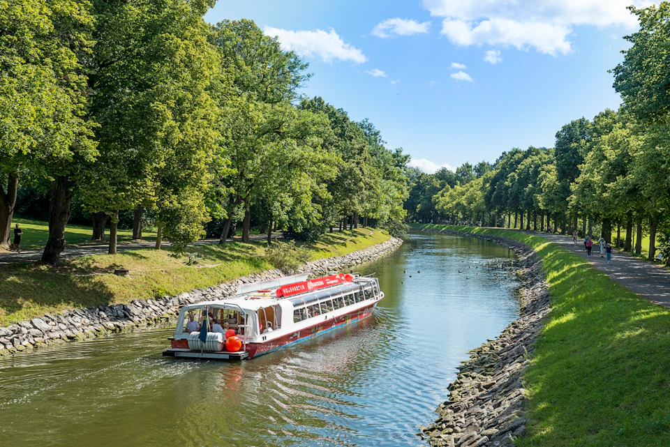 A sightseeing boat travels along a tree-lined canal under a bright blue sky with scattered clouds.
