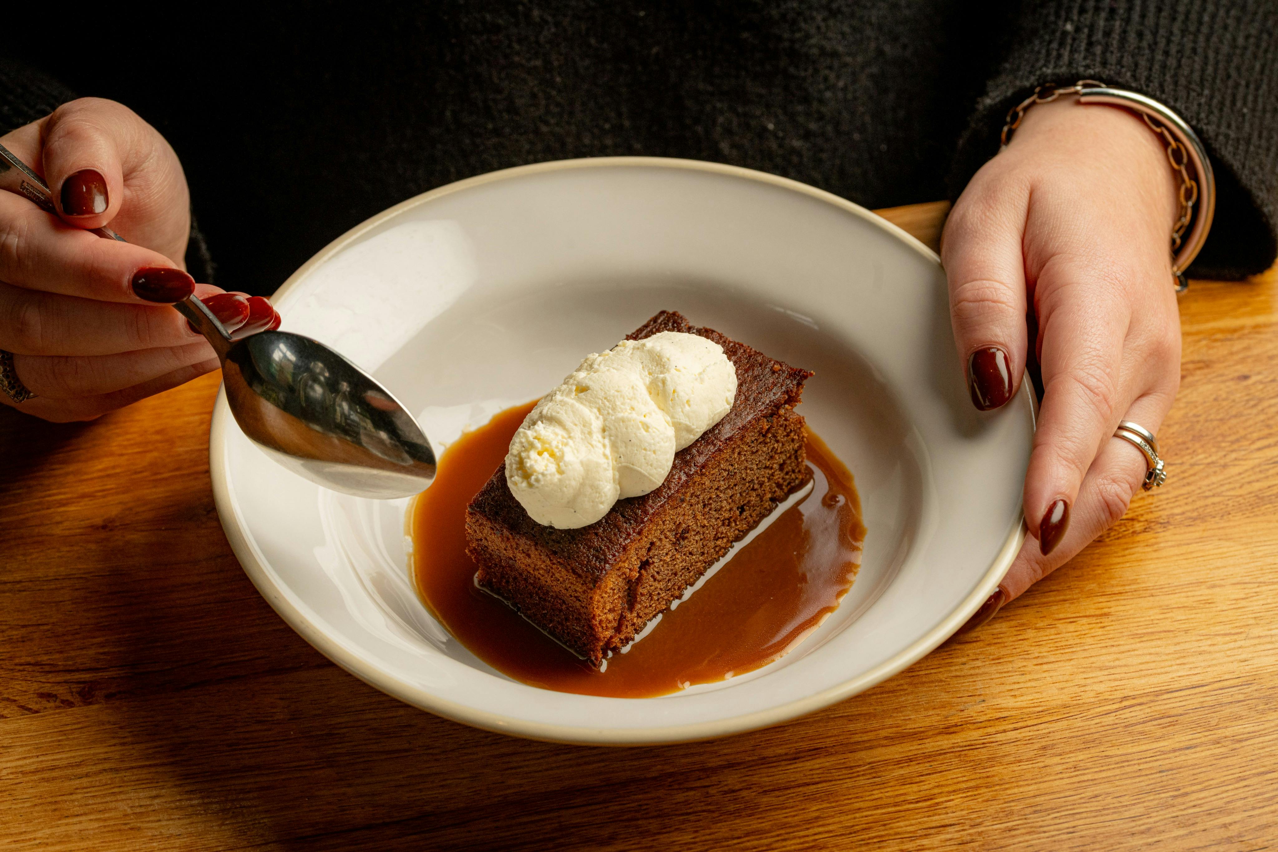 A person holding a spoon and a dish with a piece of cake topped with cream, served with caramel sauce.