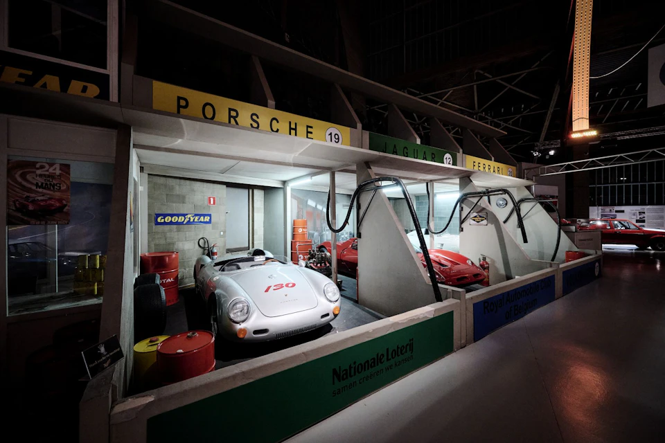 Vintage Porsche, Jaguar, and Ferrari race cars displayed in retro garage setups with tools, tires, and sponsor signage.