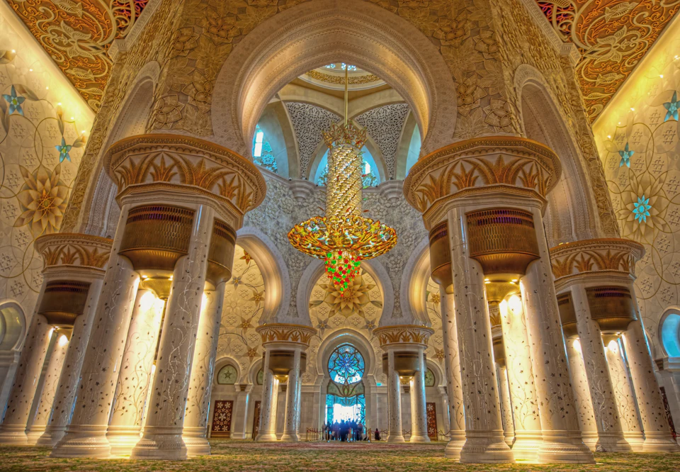 Ornate mosque interior with large golden chandelier, detailed pillars, and intricate archways. Stained glass windows in the background.