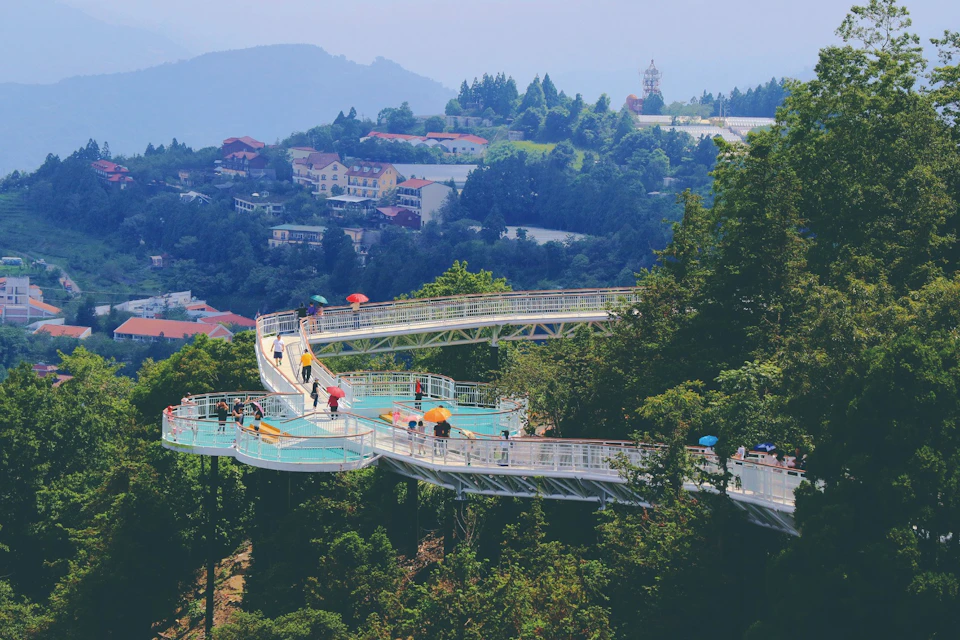 Elevated walkway in a forested area with people walking, surrounded by trees and hills with buildings in the background.