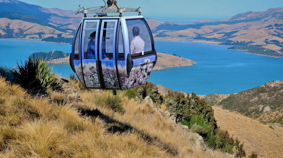 A gondola lift carrying passengers over a scenic landscape with hills, grass, and a body of water in the background.