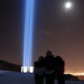 Friends hug in front of the Imagine Peace Tower at night. The moon is shining and the city of Reykjavik is in the background.
