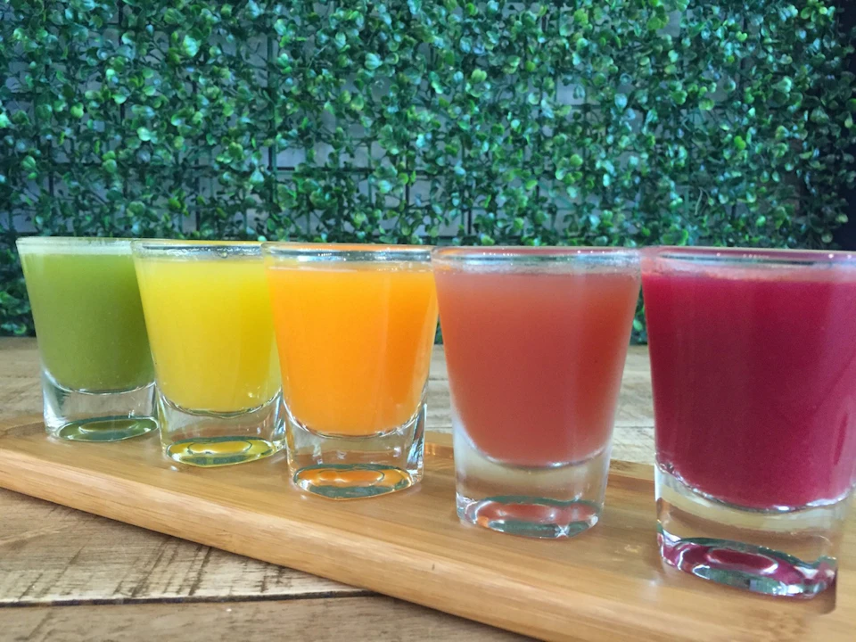 A wooden tray with five small glasses of colorful juices in green, yellow, orange, red, and purple, against a leafy backdrop.