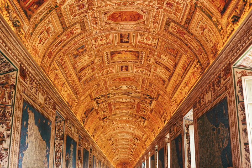 An ornate corridor with an elaborately decorated vaulted ceiling featuring intricate artwork and golden accents.