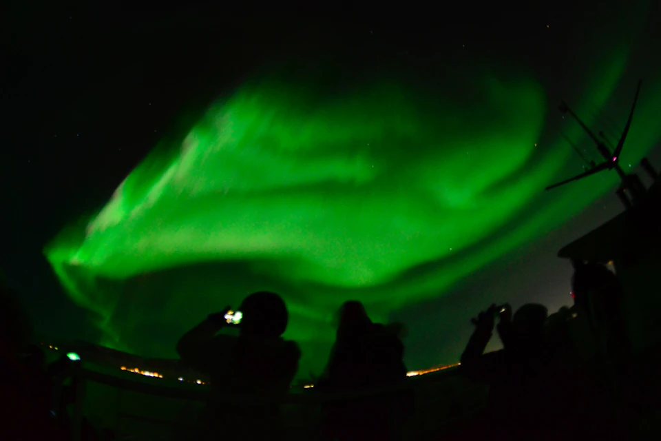 Silhouettes of passengers watching the green northern lights on a boat.