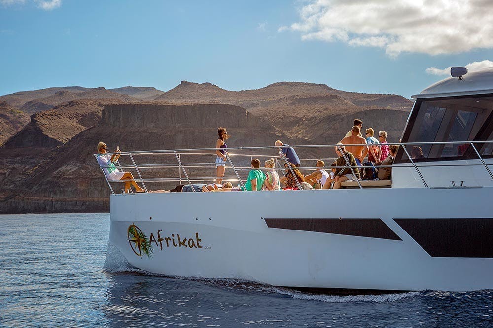 一群人乘坐一艘名为Afrikat的船，在水面上享受晴朗的一天，背景是山地。
