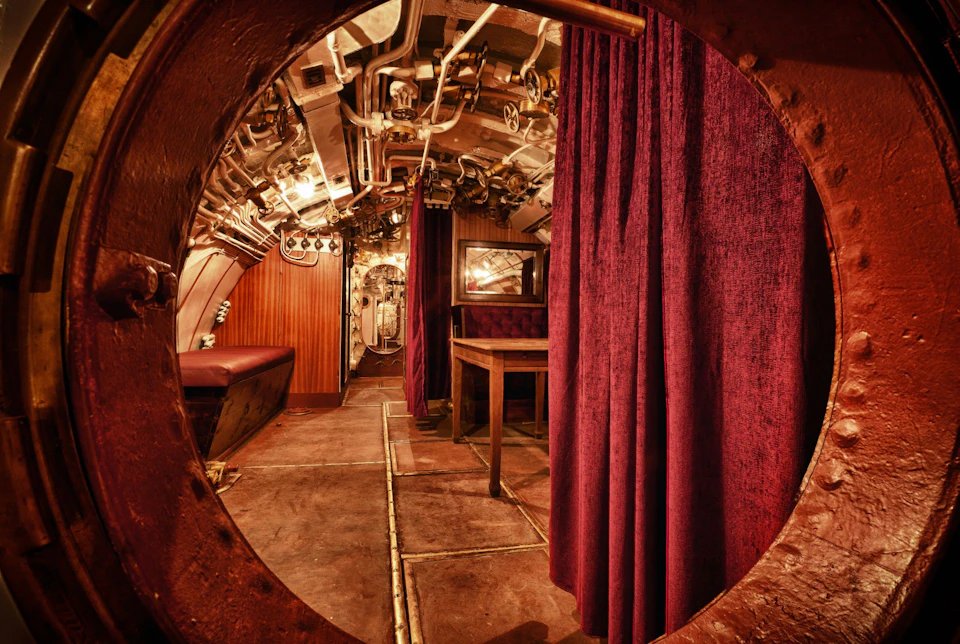 Interior of a submarine featuring a steel corridor, red curtains, exposed pipes on the ceiling, a table, and a mirror on the wall.
