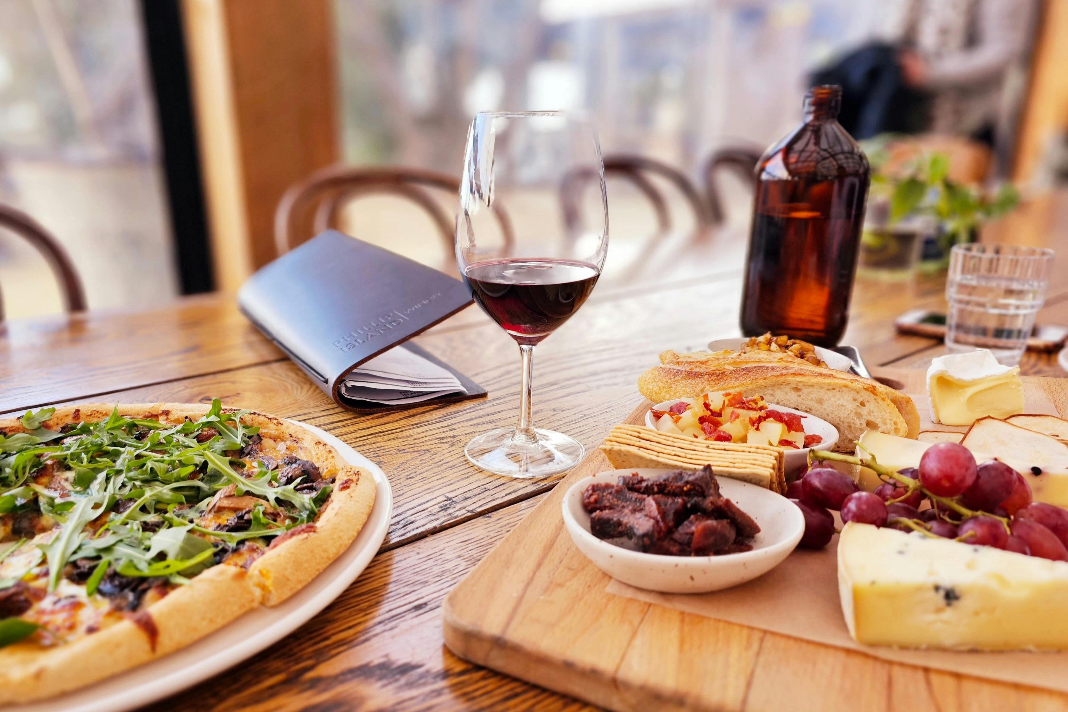 Wooden table with pizza, charcuterie board, a glass of red wine, and a brown bottle. A closed menu is also on the table.