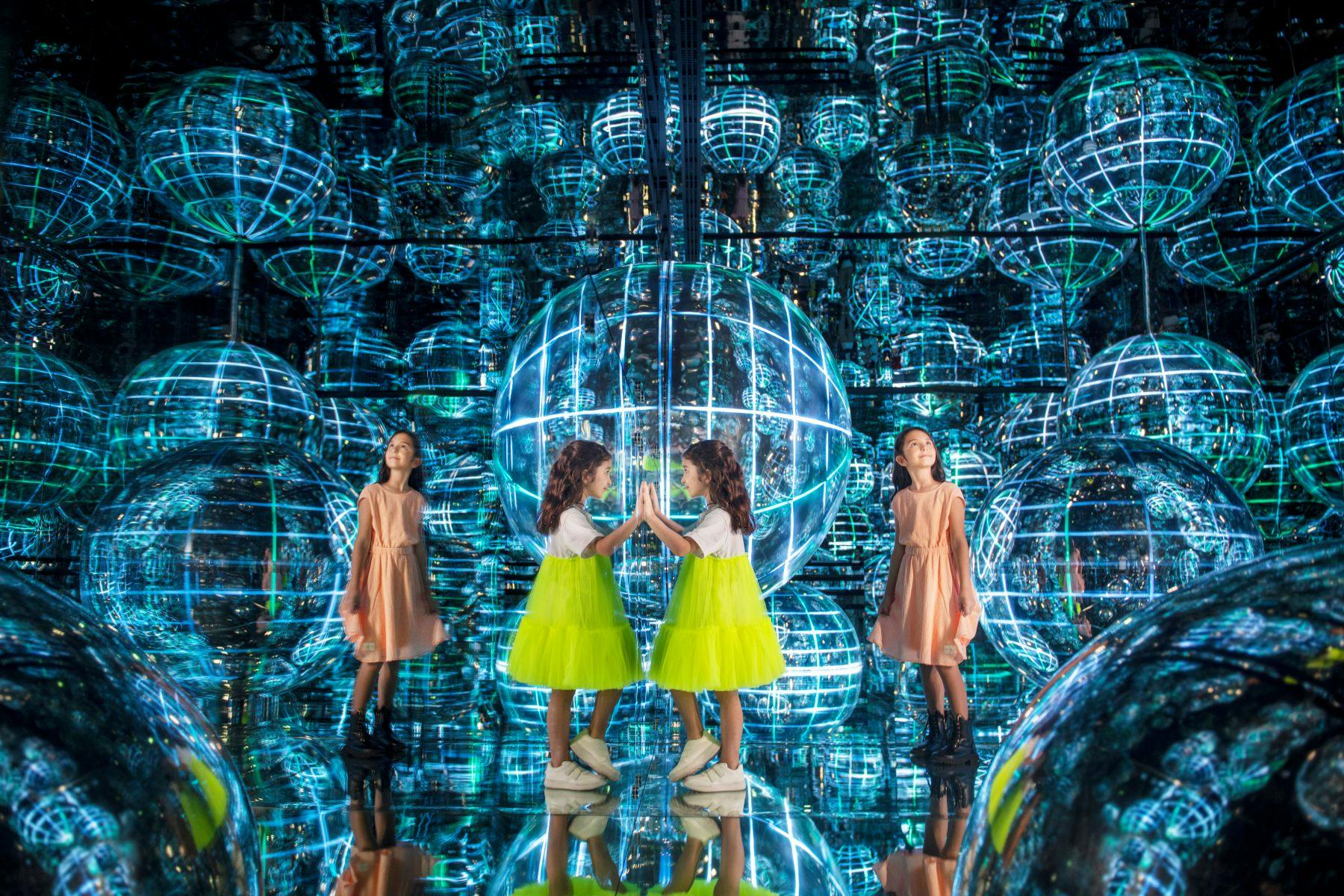 노란 치마를 입은 한 소녀가 커다란 빛나는 구가 가득한 방에서 거울을 들여다보고 있으며, 근처에 다른 소녀가 있습니다.