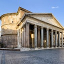 Antico Pantheon romano con grandi colonne, frontone triangolare e rotonda in una piazza vuota di ciottoli sotto un cielo limpido.