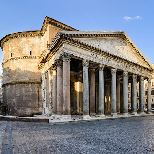Antico Pantheon romano con grandi colonne, frontone triangolare e rotonda in una piazza vuota di ciottoli sotto un cielo limpido.
