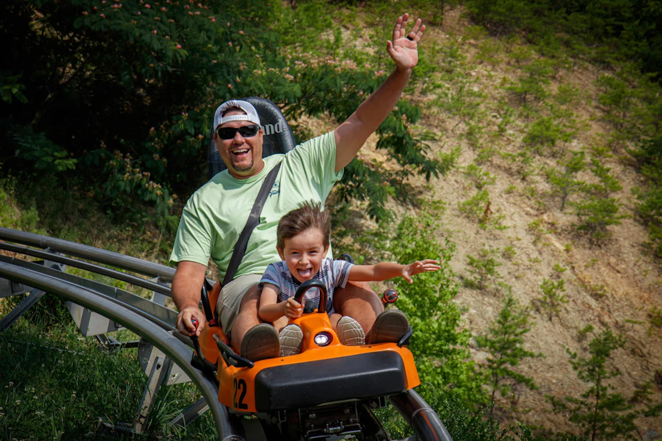 Man and child riding a roller coaster, both smiling with joy. The man has one arm raised, and greenery is visible in the background.