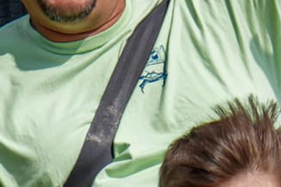 Man and child riding a roller coaster, both smiling with joy. The man has one arm raised, and greenery is visible in the background.