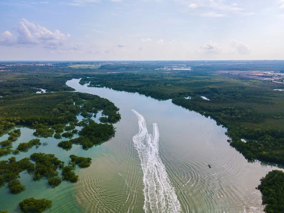 Aerial view of a river winding through green forest with boat wake patterns on the water and a clear sky above.