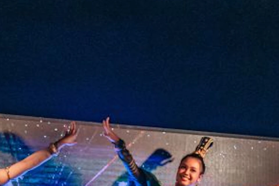 Three performers in traditional attire dance on stage, with one playing a large drum, against a vibrant digital backdrop.