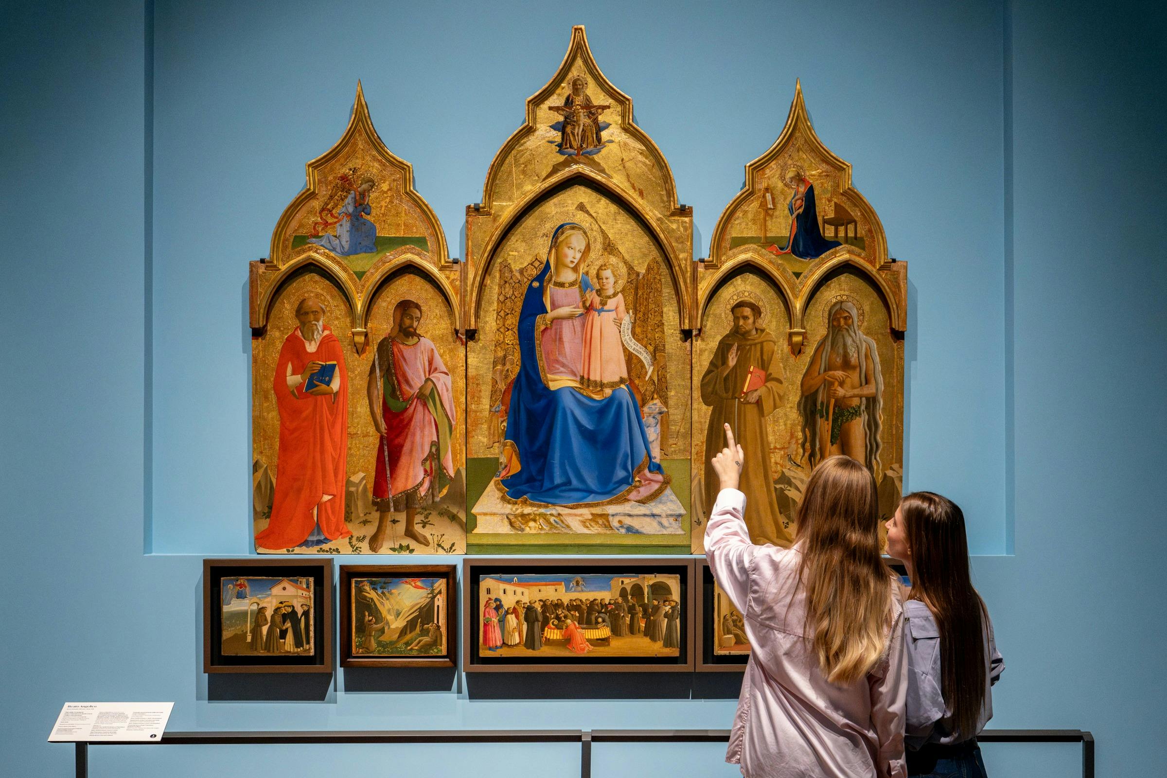 Two women stand in a museum, looking at a religious painting featuring the Virgin Mary with child and saints, framed by a gold triptych.