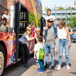 En leende kvinna ger en high-five till ett barn framför en färgglad buss. En man bär ett annat barn på axlarna bredvid en kvinna.