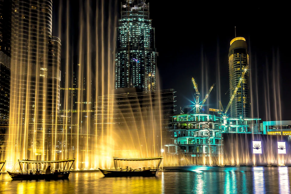 Waterfront at night with tall buildings, illuminated fountains, cranes, and boats on the water.