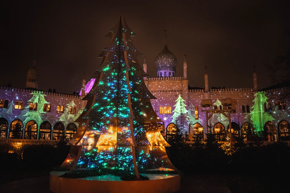 A festive illuminated tree stands in front of a building adorned with intricate lights and patterns, creating a vibrant night scene.
