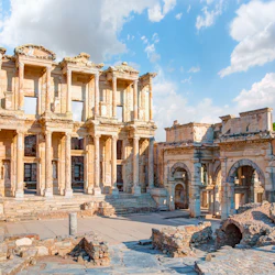 Forntida ruiner med kolonner, bågar och en stor fasad, belägna mot en ljus himmel med spridda moln.