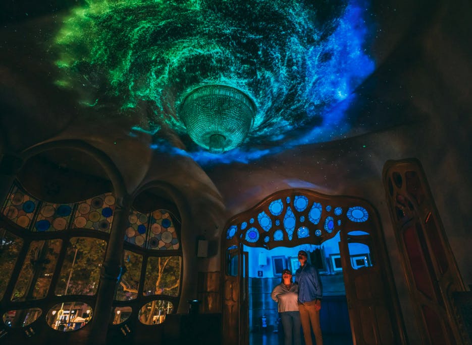 Two people stand in an ornate room with colorful stained-glass windows and a ceiling illuminated with swirling green and blue lights.